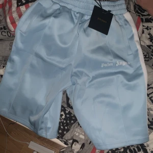 Ljusblå shorts från Palm Angels - Snygga ljusblå shorts från Palm Angels med vit logga på ena benet och vita ränder längs sidorna. Shortsen har resår i midjan och sidofickor. Perfekta för en avslappnad stil.