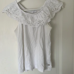 Vit blus med broderad volang från Hampton Republic - Säljer en söt vit blus från Hampton Republic med bred broderad volang upptill. Blusen är ärmlös och har fina broderade blommor på volangen. Perfekt för varma dagar och ger en romantisk känsla.