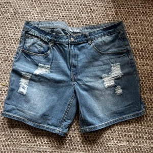 Blå jeansshorts med slitningar från Black Shrimp - Snygga blå jeansshorts från Black Shrimp med slitna detaljer framtill. Klassisk femficksmodell och normal passform. Tillverkade i 100% bomull för en skön känsla. Perfekta för varma dagar.