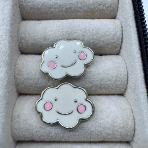 Molnformade örhängen med rosa kinder - Supergulliga örhängen i form av små vita moln med rosa kinder och ett leende ansikte. Perfekta för att ge en lekfull touch till din stil. Metallkant runt molnet och stift på baksidan.