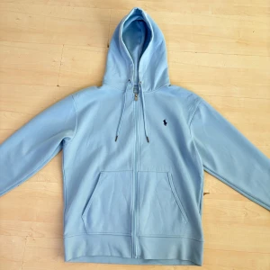 Ralph Lauren Zip - Säljer en stilren ljusblå hoodie från Ralph Lauren med dragkedja och klassisk logga på bröstet. Den har en justerbar huva och praktiska fickor framtill. Perfekt till våren och sommaren!