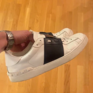 Vita Valentino Garavani sneakers - Säljer ett par vita sneakers från Valentino Garavani med bred svart rem över vristen och nitar längs sulan. Skorna har rund tå, platt sula och klassisk snörning. och boxen medföljer. Storlek 43 men tycker dom är en aning stora i storleken :), säljes då jag ej använder dom