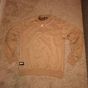 Beige sweatshirt från 1st - Säljer en beige sweatshirt från 1st med rund halsringning och tryck i vitt vid halsen. Tröjan har långa ärmar, ribbade muddar och nederkant samt en liten patch nertill. Perfekt för en avslappnad stil.
