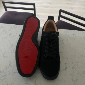Louboutin skor - Säljer ett par stilrena svarta sneakers med klassisk snörning och en unik röd sula. Skorna har rund tå och svart ovandel i mocka med beige insida. Perfekta för dig som vill sticka ut med en detalj under skon.