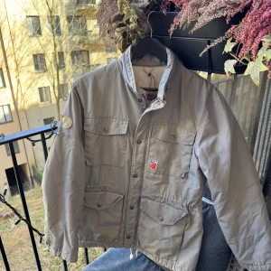 Grå field-jacka från Fjällräven - Säljer en grå jacka från Fjällräven med fyra stora fickor framtill och broderad logga på ärmen. Jackan har hög krage, dragkedja och knappar. Materialet är en blandning av polyester och bomull, med foder i nylon.