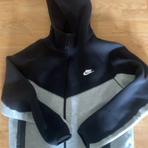 Nike tech grå svart storlek L - Säljer en snygg  Nike tech  i svart och grått med vit logga på bröstet. Den är i ritkigt bra skick allt utom att på den högra fickan så har det ena snöret gått av men det gör inget. Pris går att diskutera och hör av er vid frågor