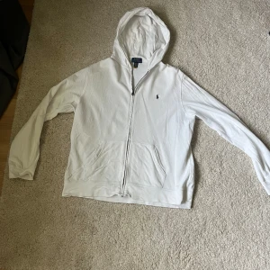 Vit hoodie från Polo Ralph Lauren - Säljer en klassisk vit hoodie från Polo Ralph Lauren i barnstorlek XL. XL i barnstorlek motsvarar XS/S i vuxenstorlek. Tröjan har dragkedja framtill, huva och två fickor. Den är dekorerad med den ikoniska broderade loggan på bröstet. Perfekt för en avslappnad och stilren look.
