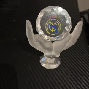 Kristallprydnad med Real Madrid-logga - Säljer en unik kristallprydnad med Real Madrid-logga. Prydnaden har två händer som håller en slipad kristall med klubbens emblem. Perfekt för fotbollsfans!