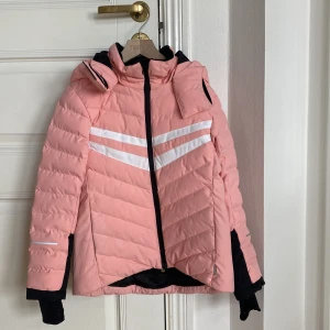 Rosa dunjacka från Reima - Säljer en aprikos skidjacka/vinterjacka  från Reima med vita ränder på bröstet och svart detalj vid ärmslut. Vattentät, vindtät och giftfritt material. Supernöjda med jackan då vi haft den i flera färger. Endast använd för skidåkning Jackan har avtagbar huva och dragkedja framtill. Perfekt för kyliga dagar och har en sportig look. Stl 134. Nypris 1199.