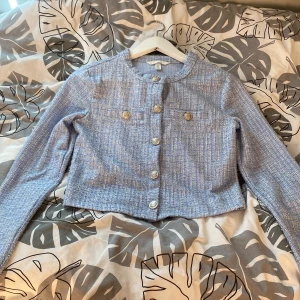 Ljusblå croppad kavajjacka från H&M - Säljer en ljusblå croppad kavajjacka från H&M med rund halsringning och dekorativa guldknappar framtill. Jackan har ett diskret vävt mönster och långa ärmar. Perfekt för att lyfta en outfit med en chic touch.