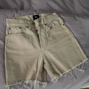 Beige jeansshorts med fransar från 157 - Säljer ett par beige jeansshorts från 157 med hög midja och råa fransade benslut. Klassisk femficksmodell med knappar framtill. Perfekta för sommaren och riktigt snygga till en enkel topp eller hoodie.