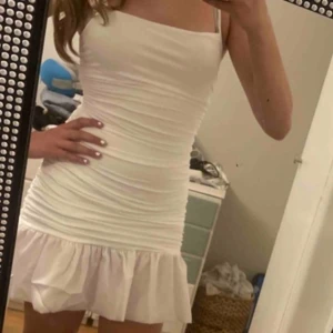 Vit kort klänning med volangkant - Säljer en söt vit kortklänning med smala axelband och figurnära passform. Klänningen har rynkade detaljer längs hela kroppen och en volangkant nertill som ger en extra touch. Perfekt för sommarens alla tillfällen!
