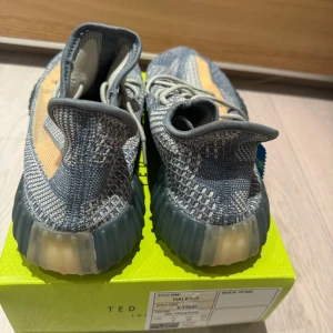 Adidas Yeezy Boost 350 V2 sneakers grå och orange - Säljer ett par Adidas Yeezy Boost 350 V2 sneakers i grått med orange sidostrip. Skorna har stickad ovandel, rund tå och platt sula. Perfekta för dig som gillar streetwear och vill ha något unikt på fötterna.