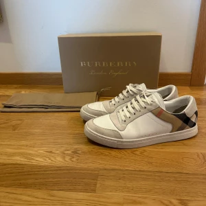 Burberry Skor - Säljer ett par snygga vita sneakers från Burberry i nytt skick. De har den klassiska Burberry-rutiga detaljen på sidan. Perfekta för både vardag och fest! Kommer med originalkartong och dustbag. Passa på att köpa dessa stilrena skor! 🏷️Storlek 41,5 passar även upp till 42,5