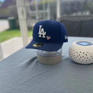 Blå LA keps från New Era - Säljer en blå keps från New Era med LA-logga framtill och broderade detaljer på sidan. Kepsen har böjd skärm och klassisk 59FIFTY-passform. Märke och storleksetikett finns kvar på insidan. Den är självklart Äkta, köpte den på hatstore.