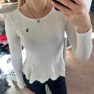 Vit långärmad topp med volang från Ralph Lauren - Säljer en vit långärmad topp från Ralph Lauren med volang nertill och rund halsringning. Toppen har en liten broderad logga på bröstet och är figurnära i modellen. 