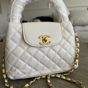Vit quiltad handväska  - Supersnygg vit handväska  med quiltat mönster och guldfärgad kedja. Väskan har ett klassiskt lock med ikonisk CC-detalj i guld och en stadig topphandtag. Perfekt för dig som vill ha en elegant accessoar med lyxig känsla. Helt ny och oanvänd 