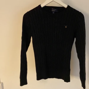 Svart kabelstickad tröja från GANT - Kabelstickad svart tröja från GANT i bomull. Klassisk rund halsringning och långärmad modell. Liten broderad logga på bröstet. Perfekt baströja med tidlös design. Pris kan diskuteras