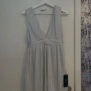 Ljusglittrig långklänning med v-ringning - Säljer en elegant ljus långklänning med djup v-ringning och bred midjedetalj. Klänningen har ett glittrigt tyg och är ärmlös, perfekt för speciella tillfällen. Materialet känns mjukt och skimrande.