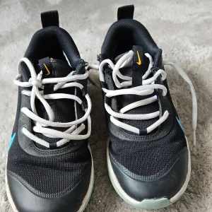 Svarta Nike sneakers med blå detalj. - Säljer ett par svarta Nike sneakers med vit sula och blå swoosh på sidan. Skorna har vita snören och en svart ovandel i syntetmaterial. Bak på hälen finns en blå blomdetalj. Perfekta för vardagsbruk.