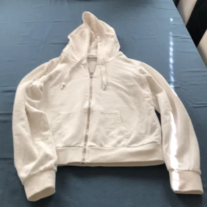 Vit hoodie med dragkedja från Anna Field - Säljer en klassisk vit hoodie från Anna Field med dragkedja framtill och huva med snörning. Tröjan har långa ärmar, ribbade muddar och två fickor framtill. Perfekt för en avslappnad stil.