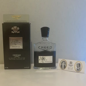 Creed Aventus Eau de Parfum 100ml - Lyxig parfym från Creed, Aventus, i en elegant genomskinlig glasflaska med svart etikett och silverdetaljer. Flaskan rymmer 100 ml och har en svart kork. Doften är ikonisk och exklusiv, perfekt för dig som vill sticka ut.