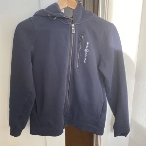 Mörkblå hoodiejacka från Sail Racing - Mörkblå hoodiejacka från Sail Racing med dragkedja framtill och på bröstfickan. Jackan har huva och logga tryckt på bröstet. Perfekt för dig som gillar sportig stil och vill ha något bekvämt och snyggt.