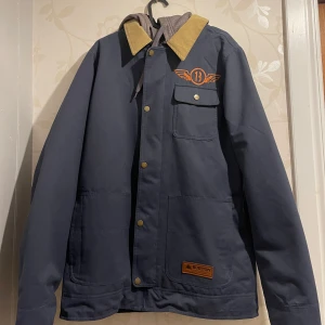 Blå jacka från Burton med huva - Säljer en blå vinterjacka från Burton med beige krage och grå huva. Jackan har tryckknappar, dragkedja, bröstficka med lock och orange broderad logga på bröstet samt stort tryck på ryggen. 