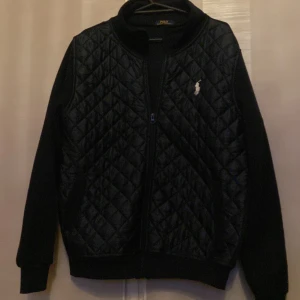 Svart quiltad jacka från Polo Ralph Lauren - Säljer en svart quiltad jacka från Polo Ralph Lauren med dragkedja framtill och broderad logga på bröstet. Jackan har långärmade ärmar och ribbade muddar. Perfekt för kyliga dagar.
