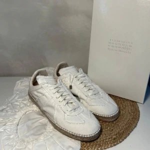 Maison Margiela vita sneakers - Maison Margiela sneakers i helvitt med unik, rå kantdesign och synliga sömmar. Skorna har snörning och en beige sula med klassiskt mönster undertill. Medföljer vita skyddande skopåsar och originalkartong. Perfekta för dig som gillar stilrena och trendiga detaljer.helt nya allt og