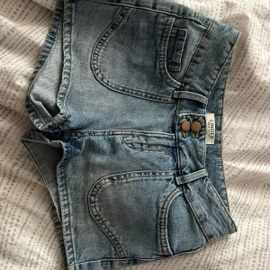 Blå jeansshorts från Lindex - Snygga blå jeansshorts från Lindex med dubbla knappar framtill och dekorativa broderade hjärtan på bakfickorna. Använda två gånger och jag säljer dem pga att de är för små! Klassisk femficksmodell och låg midja. Perfekta för sommaren!