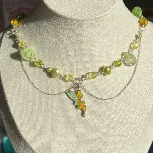 Handgjort halsband med Tingeling-berlock  - Unikt halsband med gröna och gula pärlor i olika former, dekorerat med en söt Tingeling-berlock i mitten. Kedjan är silverfärgad och har dubbla lager framtill för extra detalj. Perfekt för dig som älskar sagor och färgglada accessoarer.