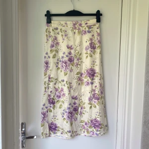 Vit blommig långkjol med lila rosor - Supersöt långkjol med blommigt mönster i lila och grönt på vit botten. Kjolen har en rak passform och resår i midjan för extra komfort. Perfekt för en somrig och romantisk look.