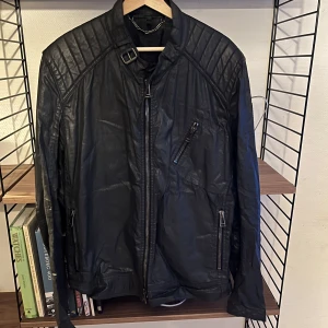Svart vårjacka från Belstaff - Säljer en svart vårjacka från Belstaff med coola quiltade axlar och flera dragkedjor. Jackan har ståkrage med spänne och snygga detaljer på ärmarna. Perfekt för dig som gillar en klassisk bikerstil.