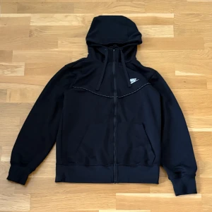 Nike Hoodie  - Svart hoodie från Nike med hel dragkedja och huva. Klassisk logga på bröstet och snörning i huvan. Perfekt för en sportig stil eller till vardags. Mjuk och bekväm med normal passform.