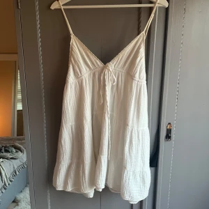 Linen vit somrig klänning med smala axelband - Söt vit miniklänning med smala axelband och knytband framtill. Lätt och luftig design med volang nertill, perfekt för varma dagar.