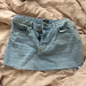 Ljusblå jeanskjol med rå kant - Säljer en ljusblå jeanskjol från Nelly med klassisk femficksdesign och rå, fransig nederkant. Kjolen har bälteshällor och knappgylf. 