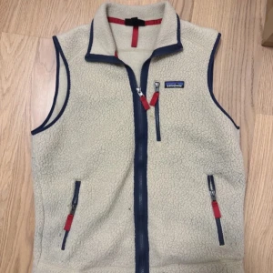 Beige fleeceväst från Patagonia - Säljer en beige fleeceväst från Patagonia med blå detaljer och röda dragkedjor. Västen har hög krage, tre fickor med dragkedja och klassisk Patagonia-logga på bröstet. Perfekt för lager-på-lager och outdoor-stil. Storlek M passar bra runt 190-185 på längden. Hör av er vid funderingar😄