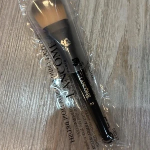 Lancôme Mini Foundation Brush - En mini foundationborste från Lancôme med svart handtag och täta, mjuka borst i beige och svart. Perfekt för applicering av flytande foundation. Kommer i originalförpackning och är smidig att ta med i necessären.