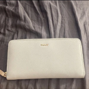 Ljusgrå plånbok från DKNY - Säljer en stilren ljusgrå plånbok från DKNY med guldfärgad dragkedja. Plånboken har flera fack för kort och mynt, samt ett innerfack med dragkedja. Perfekt för att hålla ordning på dina värdesaker.