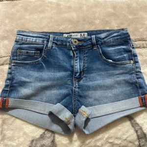 Blå jeansshorts från FB Sister - Snygga blå jeansshorts från FB Sister med klassisk femficksdesign och uppvikta benslut. Shortsen har normal passform, knapp och dragkedja framtill samt bälteshällor. Perfekta för varma dagar och enkel att matcha med olika toppar.