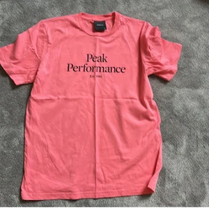 Rosa t-shirt från Peak Performance - Säljer en rosa t-shirt från Peak Performance med svart tryck på bröstet. Klassisk passform och rund halsringning. Perfekt för en avslappnad stil och enkel att matcha med jeans eller shorts.