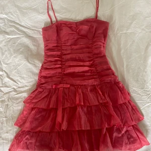 Rosa volangklänning med smala axelband - Säljer en söt rosa kortklänning med smala axelband och flera lager volanger. Klänningen har rynkade detaljer framtill och en liten rosett i midjan. Perfekt för dig som vill sticka ut med en färgglad och lekfull look.