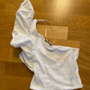 Vit oneshoulder topp med volang - Säljer en vit oneshoulder topp med en bred volang på ena axeln och ett smalt axelband på andra sidan. Toppen är i ett mjukt material och har en trendig asymmetrisk design.
