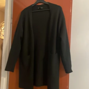 Svart lång cardigan kofta  - Säljer en svart lång cardigan med öppen front och två fickor framtill. Koftan har lång ärm och en rak, avslappnad passform. Perfekt att slänga över en t-shirt eller tröja för en enkel och stilren look.
