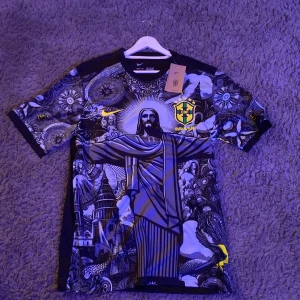 Brasilien fotbollströja med Christ The Redeemer -tryck från Nike - Helt oanvänd Brasilien fotbollströja med stort tryck av Kristusstatyn och detaljerade grafiska mönster. Tröjan har korta ärmar, svart bas med inslag av gult och blått samt ett sytt Brasilien-emblem på bröstet.