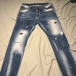Dsquared2 slitna blå jeans - Säljer ett par blå jeans från Dsquared2 med slitningar och hål för en riktigt edgy look. Jeansen har klassisk femficksdesign, gylf med dragkedja och raka ben. Perfekta för dig som gillar streetstyle och vill sticka ut.
