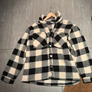 Svart och vit rutig overshirt från Lindex 170 (UK14Y) - Säljer en svart och vit rutig overshirt från Lindex med klassisk krage och knappar framtill. Jackan har två stora bröstfickor med lock och långa ärmar. Perfekt för lager-på-lager och en snygg streetstil. Den är använd ett fåtal gånger men nytt skick. Den är i strl 170 (UK14Y). Det är bara att höra av sig vid närmaste fundering 😃🙌