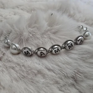 Armband med bokstäver 'MAMMA' och pärlor - Snyggt tennarmband med silverfärgade bokstavspärlor som bildar ordet 'MAMMA' och vita pärlor. Armbandet har en tunn silverfärgad kedja och justerbart lås. Perfekt som present till någon speciell. Handledsmått 17,5-22cm. 