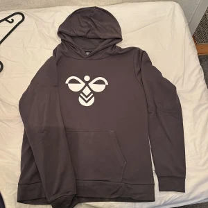 Mörkgrå hoodie från Hummel - Säljer en mörkgrå hoodie från Hummel med deras klassiska logga i vitt på bröstet. Tröjan har huva, lång ärm och en stor magficka framtill. Perfekt för en sportig och avslappnad stil.